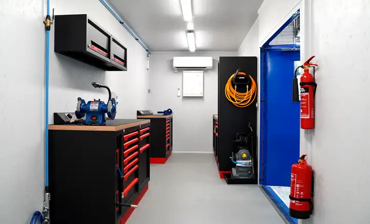Interior of a fully equipped shipping container workshop featuring fitted tool cabinets, workbenches with bench vices, overhead storage, air conditioning unit, safety equipment, and power tools, with a brightly painted entry door.