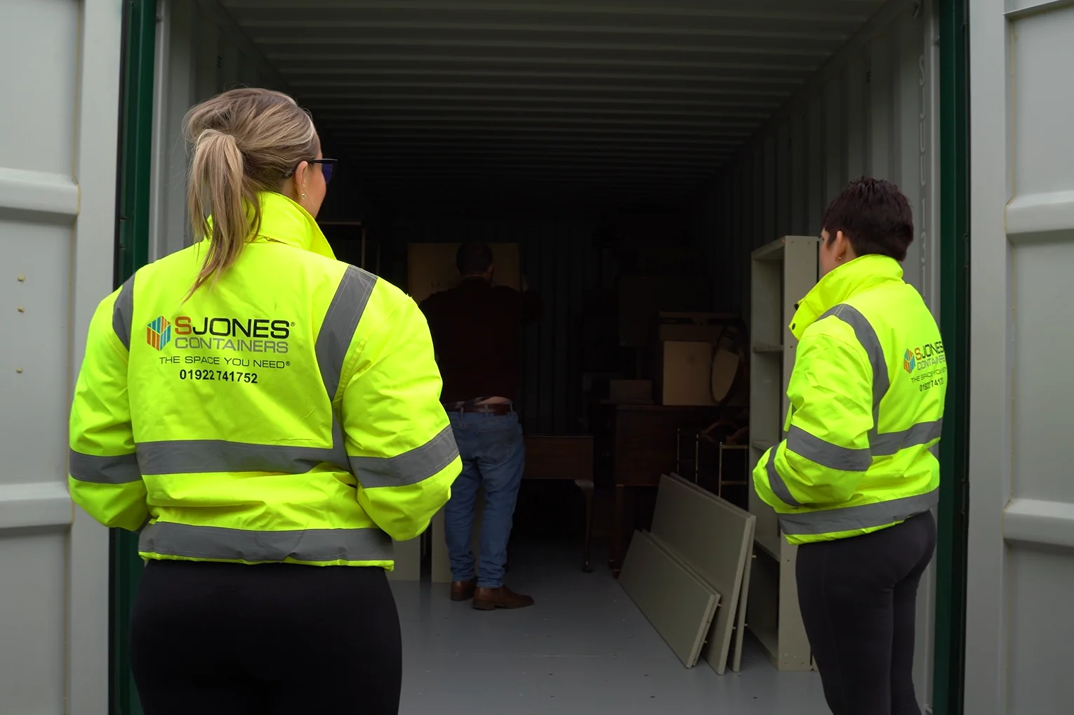 Two individuals in high-visibility yellow jackets with reflective stripes are standing at the entrance of an open storage container, looking inside. The jackets display the logo for 