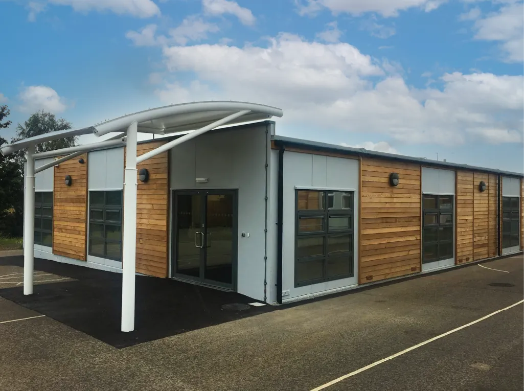 A contemporary modular building with wooden paneling and large glass windows. A white canopy supported by metal poles extends over the entrance, providing shelter. The paved area in front suggests a commercial or educational use.