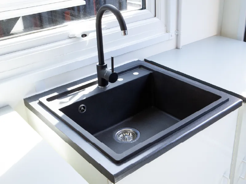 Close-up of a modern black composite sink with a matching black tap, set into a white worktop beneath a bright window inside a converted container.