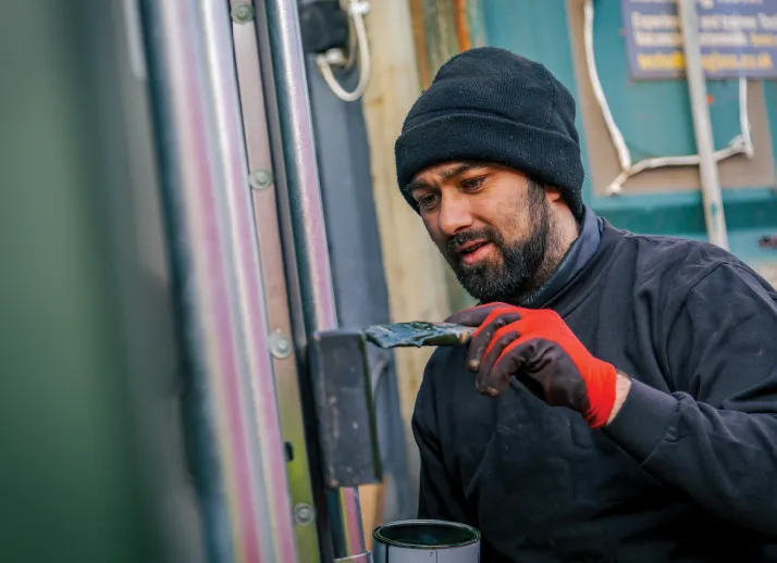 A man in a black beanie and gloves carefully applies paint to the metal handle of a shipping container, focusing on detailed restoration work.