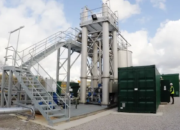 A water treatment installation with large vertical tanks and a staircase, with green containerised units helping operation. A worker in PPE visible on the right.