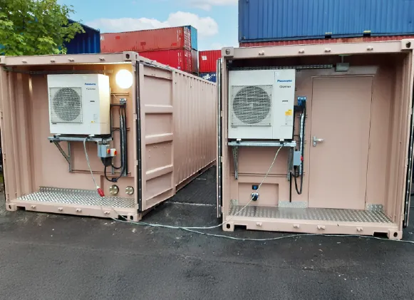Two modified beige shipping containers fitted with Panasonic air conditioning units on the exterior. One unit has a personnel door on the right side, while both containers feature elevated platforms and electrical connections.