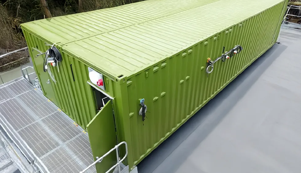 Elevated view of a large green converted shipping container by S Jones Containers, installed on a raised platform with secure railings. Outfitted with external fittings, cabling, and access doors, the container is configured for use as a switchgear enclosure on the HS2 infrastructure project site.