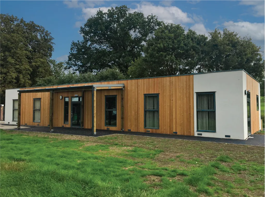 A modular building with a mix of wooden and white paneling, surrounded by greenery. Large windows allow natural light inside, and the entrance features a covered walkway supported by wooden beams.
