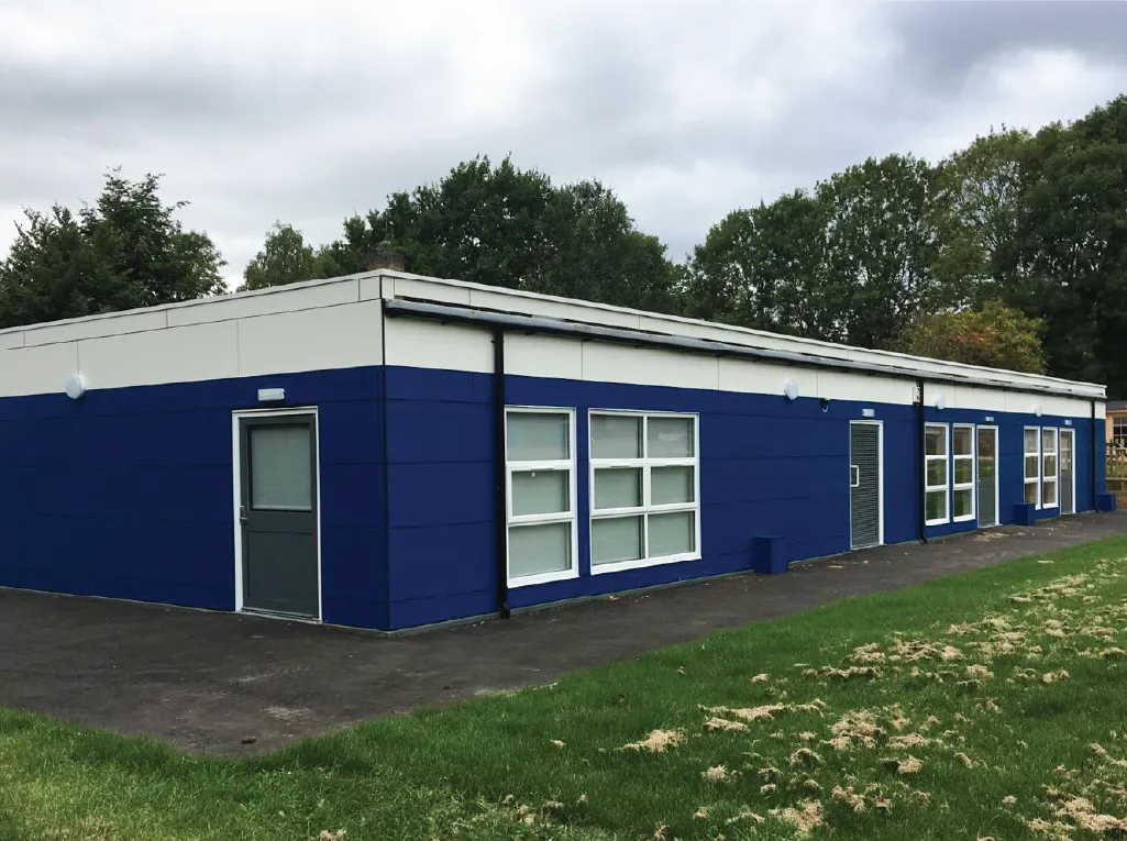 A single-story modular building with a bold blue and white exterior, multiple windows, and a grey entrance door. The structure is surrounded by a grassy area and trees, with a paved walkway leading to the entrance.