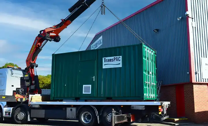 A green shipping container labeled "TransPAC" is being unloaded from a flatbed truck using a large red hydraulic crane. The container is equipped with a side personnel door and a ventilation panel. The scene takes place outside a warehouse with corrugated metal siding and red trim, under a clear blue sky.