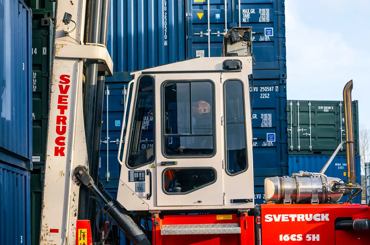A close-up of a Svetruck container handler in operation at the S Jones Containers depot in Aldridge. The cab of the handler is in focus, showing a bald-headed operator inside, concentrating on maneuvering the machine. The large lifting mechanism is visible on the left side, with stacked blue and green shipping containers forming the industrial background. The red body of the vehicle prominently displays the 'Svetruck' branding and model number '16CS 5H'. The image captures the heavy-duty machinery in action within the depot environment.