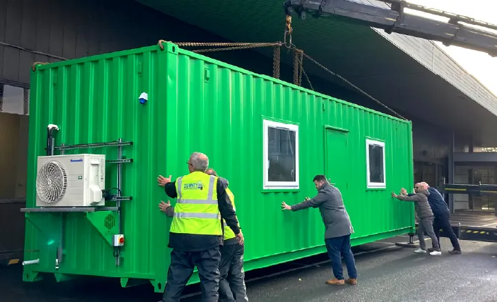 A bright green converted shipping container being maneuvered into place with the help of several people, using a crane. The unit has windows, a door, and an external air conditioning unit.