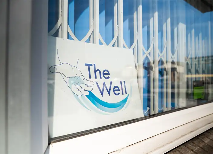A sign for “The Well” foodbank displayed in a window in Wolverhampton. The sign features an illustrated hand cupping a flowing wave beneath the organisation’s name, symbolising care and support. The window has a white security grille and reflects the surrounding street.