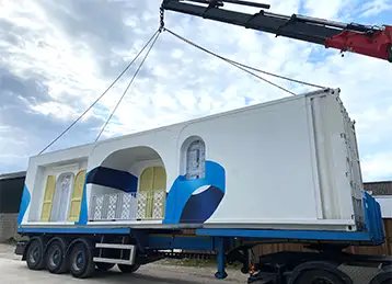 A decorated shipping container for the House of Peroni being lifted by crane from a flatbed lorry. The container is painted white with artistic blue accents and features architectural detailing including arched windows, doors, and a small balcony area, giving it a Mediterranean-style aesthetic. The scene takes place outdoors under a partly cloudy sky.