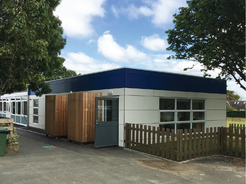 A modular school building with a white and blue exterior, featuring large windows and a grey entrance door. A small wooden fence and wooden-clad utility enclosures are positioned near the entrance, with trees and a playground visible in the background.