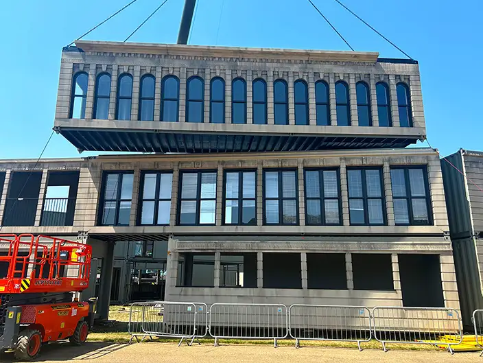 A large shipping container unit styled to resemble a traditional stone building façade is being lifted by crane and positioned on top of a two-storey modular structure. The installation, part of the Flat Iron venue, features rows of arched and rectangular windows, realistic architectural detailing, and is constructed using containers supplied by S Jones Containers. A red cherry picker and safety barriers are visible on-site during the build.