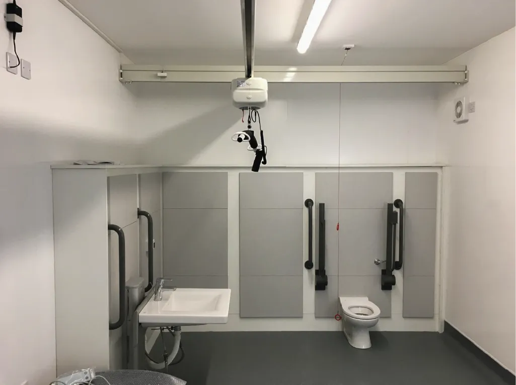 A spacious, accessible restroom inside a modular building, featuring a sink, toilet, grab rails, and a ceiling-mounted hoist system for mobility assistance. The walls are painted white with light grey paneling.