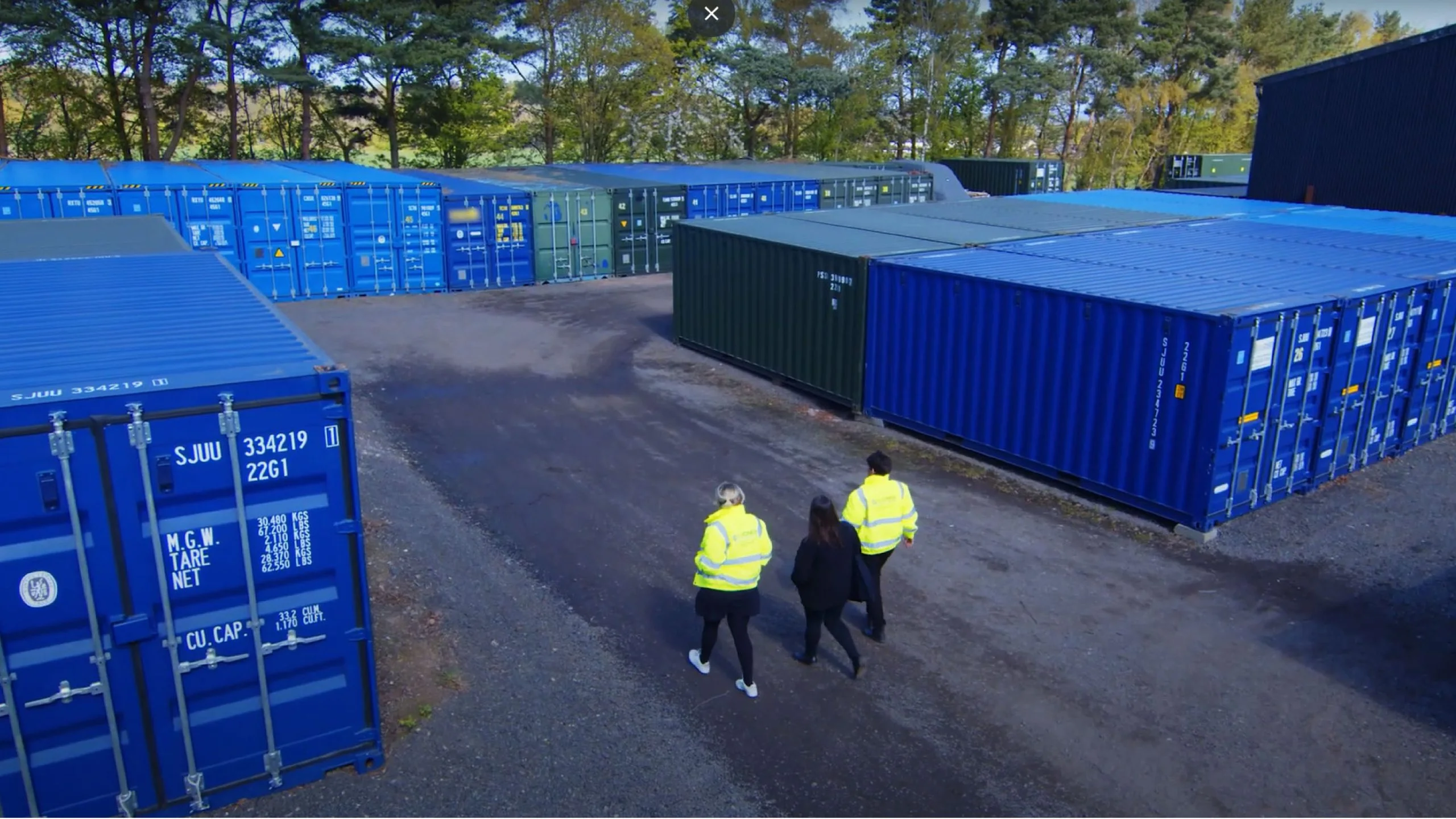 An outdoor self-storage facility with multiple shipping container units in blue and green, neatly arranged on a paved lot. Three people, two wearing high-visibility yellow jackets and one in black clothing, walk along a pathway through the storage area. The facility is surrounded by trees and greenery, with a mix of open and closed containers visible. The sky is clear, and the setting appears to be in a semi-rural or industrial location.