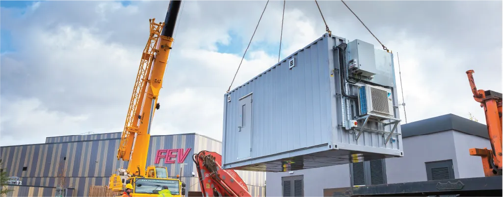 A battery storage container conversion unit being lifted by a crane at Coventry University. The unit is equipped with external air conditioning and electrical installations. The background features a large industrial building with the letters &quot;FEV&quot; visible.