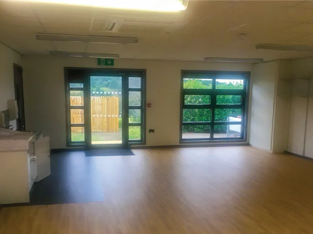 The interior of a modular building with an open-plan layout, wooden flooring, and large windows providing natural light. A set of glass doors leads outside to a fenced area with greenery. The room suggests it is a classroom space.