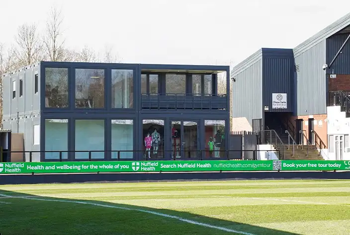 A two-storey modular hospitality suite made from converted shipping containers with large glass windows and a balcony, situated next to a sports pitch and main building, with people visible inside. A Nuffield Health advertising board runs along the edge of the field.