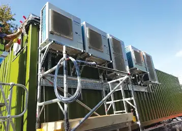Multiple external air conditioning units mounted on a green converted shipping container, supported by a metal scaffold structure. Electrical cables and conduits are connected to the units, and the setup is part of an industrial installation under a clear blue sky.