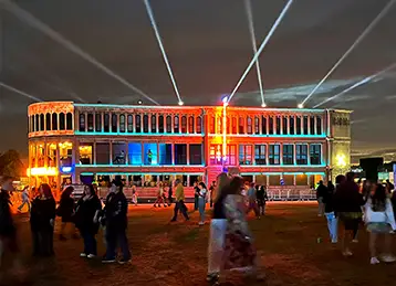 The Flatiron Central event structure, built from modified shipping containers, is illuminated with vibrant lighting effects at night. The façade, designed to resemble a classical stone building, is enhanced by beams of light projected into the sky. Event attendees walk and gather on the grass in front of the building, creating a lively festival atmosphere.