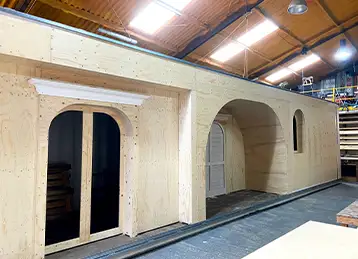 A partially constructed timber façade section inside a workshop, forming part of the House of Peroni installation. The structure features arched doorways and windows with classical-style moulding details, being prepared for attachment to a container-based build. The setting includes high ceilings, industrial lighting, and visible tools and materials, indicating an active fabrication environment.