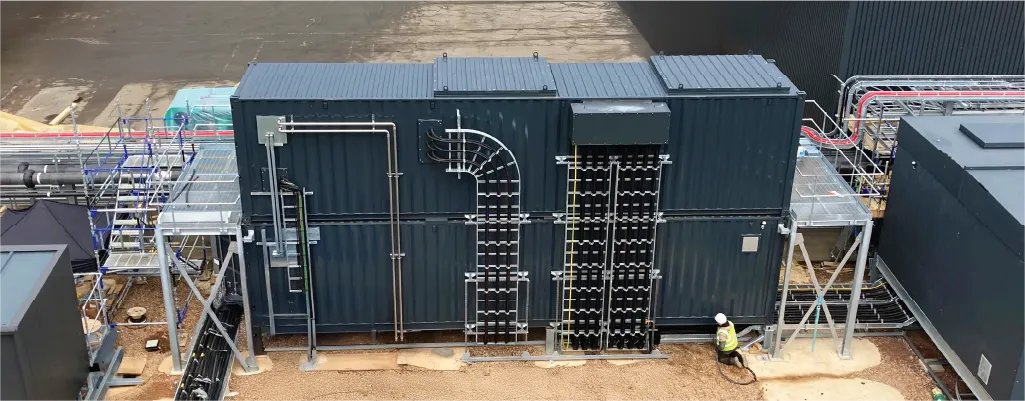 A large industrial container conversion unit with various external electrical and piping installations. A worker in a high-visibility vest is seen working near the base, and metal scaffolding and cable management systems surround the unit.