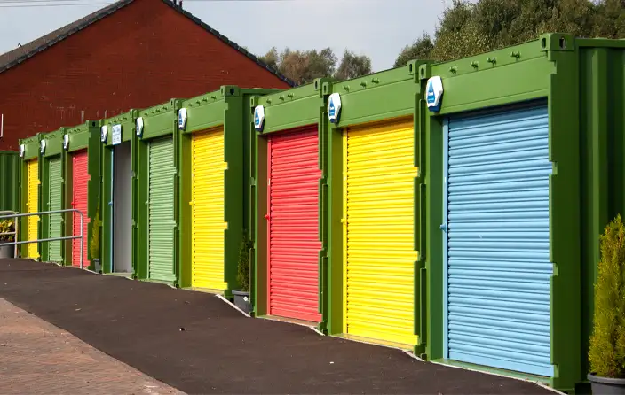 Row of converted green shipping containers with brightly coloured roller shutter doors in blue, yellow, red, and green, used as secure storage units or workshops on a tarmac path next to a brick building.