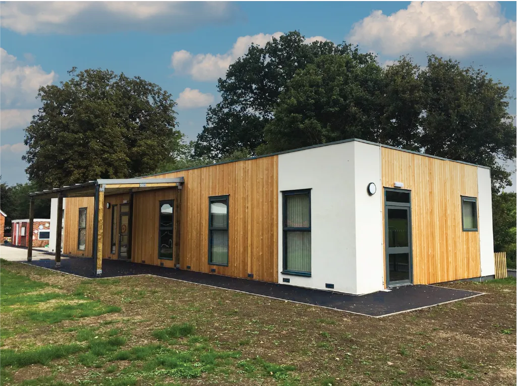 A modern modular building with a wooden and white exterior, large windows, and a covered entrance. The building is set against a backdrop of green trees and a blue sky with scattered clouds.