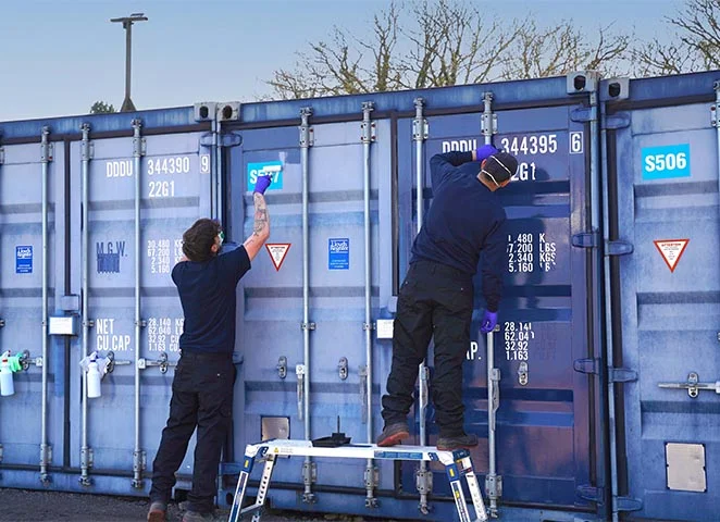 Two workers in dark uniforms are repainting identification markings on blue shipping container doors. One stands on a short step ladder while the other reaches up from the ground. Spray bottles hang from the door handles, and both workers are using small paint rollers. Trees and a lamppost are visible in the background.