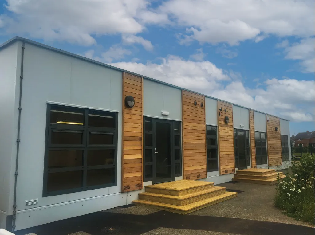 A modern modular building with a combination of white panels and wooden cladding, featuring large windows and multiple entrances. Wooden steps lead up to each entrance, and the sky above is partly cloudy.