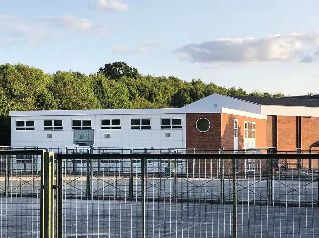 A school or community center with a modular extension featuring white and red brick walls, large windows, and a circular window near the entrance. The building is enclosed by a metal fence, with trees in the background under a clear sky.