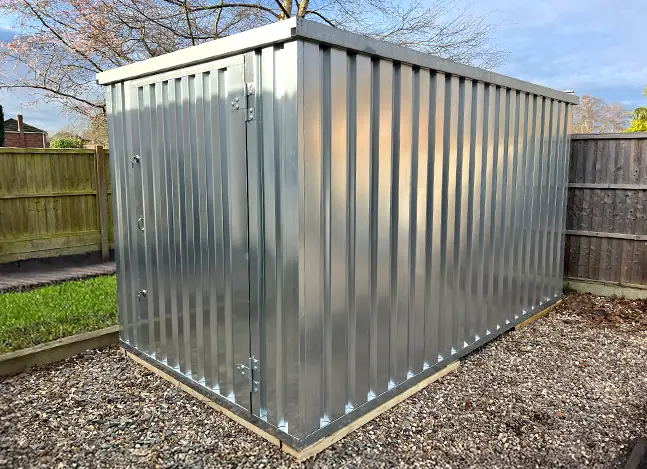 A silver flat pack metal storage shed assembled in a back garden, placed on a gravel base and positioned against wooden fencing.