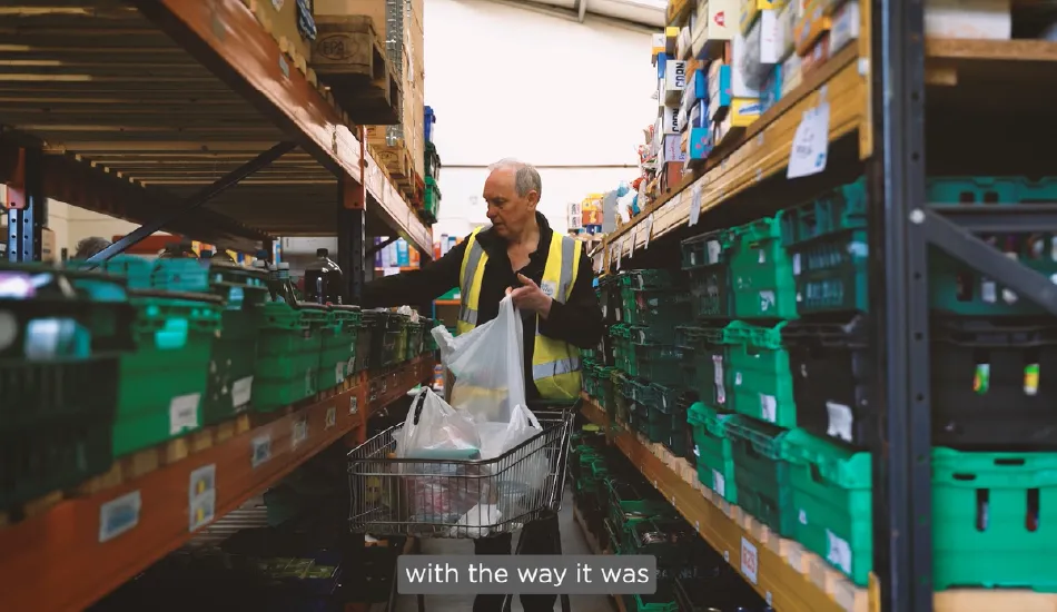 A man wearing a yellow safety vest is packing groceries into plastic bags in a warehouse aisle lined with green storage crates on metal shelves. He is placing the bags into a shopping cart. The environment resembles a food bank or supply distribution center.