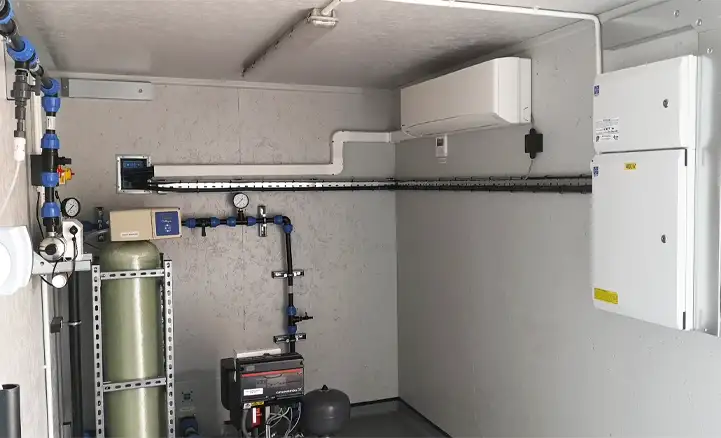 Interior of a containerised water treatment unit, featuring a vertical pressure vessel, various blue and black pipes with gauges and valves, a control panel on the wall, and an air conditioning unit for climate control. The setup is clean and organized, with all equipment mounted securely against the light grey walls.