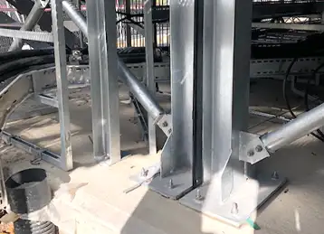 Close-up view of steel support columns bolted to a concrete base, forming part of the structural framework for a containerised installation. Electrical cables and metal conduit trays are visible in the background, indicating an industrial or infrastructure setting.