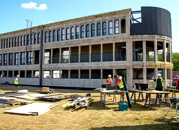 Construction underway on the Flatiron Central event structure, built from modified shipping containers with classical-style façades. The curved front section is partially clad, while workers in high-visibility gear operate on the ground, surrounded by tools, timber, and building materials. The setup takes place on a grassy area under clear skies.