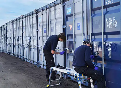 Two workers from S Jones Containers wearing protective gloves and masks apply restoration treatment to a row of shipping containers, using brushes and rollers. The containers show varying degrees of colour fade.
