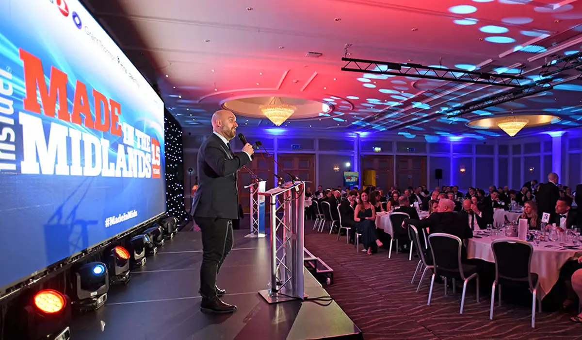 A lively scene from the Made in the Midlands Awards 2025, showing a speaker on stage addressing a large seated audience in a ballroom. The speaker, dressed in a suit, stands at a podium in front of a large digital screen displaying the event's branding. The room is filled with round banquet tables, elegantly set and surrounded by guests in formal evening attire. The ceiling is lit with vibrant red and blue lighting effects, and chandeliers add a touch of elegance to the venue.