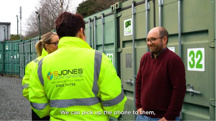 A group of people are having a conversation in front of green shipping containers with numbered labels. Two individuals are wearing high-visibility yellow jackets with 'S Jones Containers' branding, while a man in a burgundy sweater and glasses stands opposite them, smiling. The setting appears to be an outdoor storage facility with gravel ground.