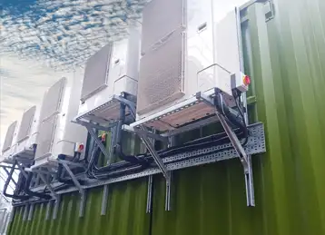 A row of large external HVAC units securely mounted to the side of a green converted shipping container. The units are supported by a robust metal framework with cable trays and conduits for power and control connections, set against a cloudy sky.