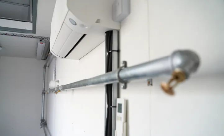 Close-up of an internal wall-mounted air conditioning unit inside a container conversion, with exposed metal pipework, sprinklers and wiring.