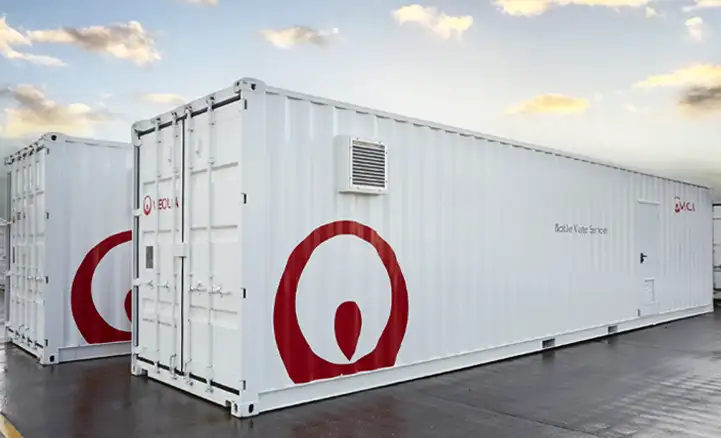 Two white shipping containers branded with the Veolia logo are positioned outdoors on a wet tarmac surface. The containers have external ventilation units and are labeled "Mobile Water Services." The sky is partly cloudy with soft light from a setting or rising sun.