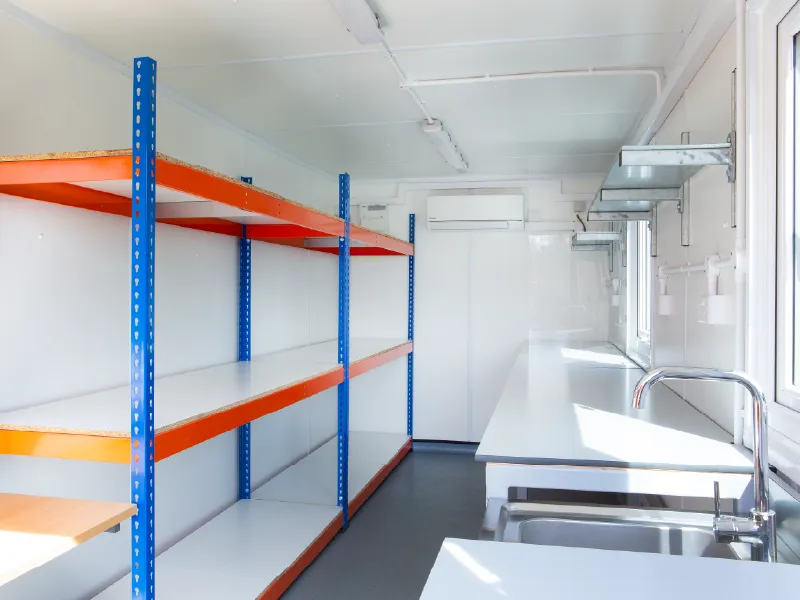 Inside view of a container lab or workshop, featuring open blue and orange industrial shelving on the left and long white worktops with metal shelves and wall sockets on the right.