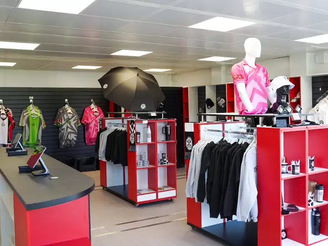 Interior of the Hednesford Town Football Club merchandise shop, featuring neatly arranged red and black display units with football apparel, hoodies, umbrellas, mugs, and other branded merchandise. A mannequin dressed in a pink team shirt stands on a central display, and the walls showcase various football kits.