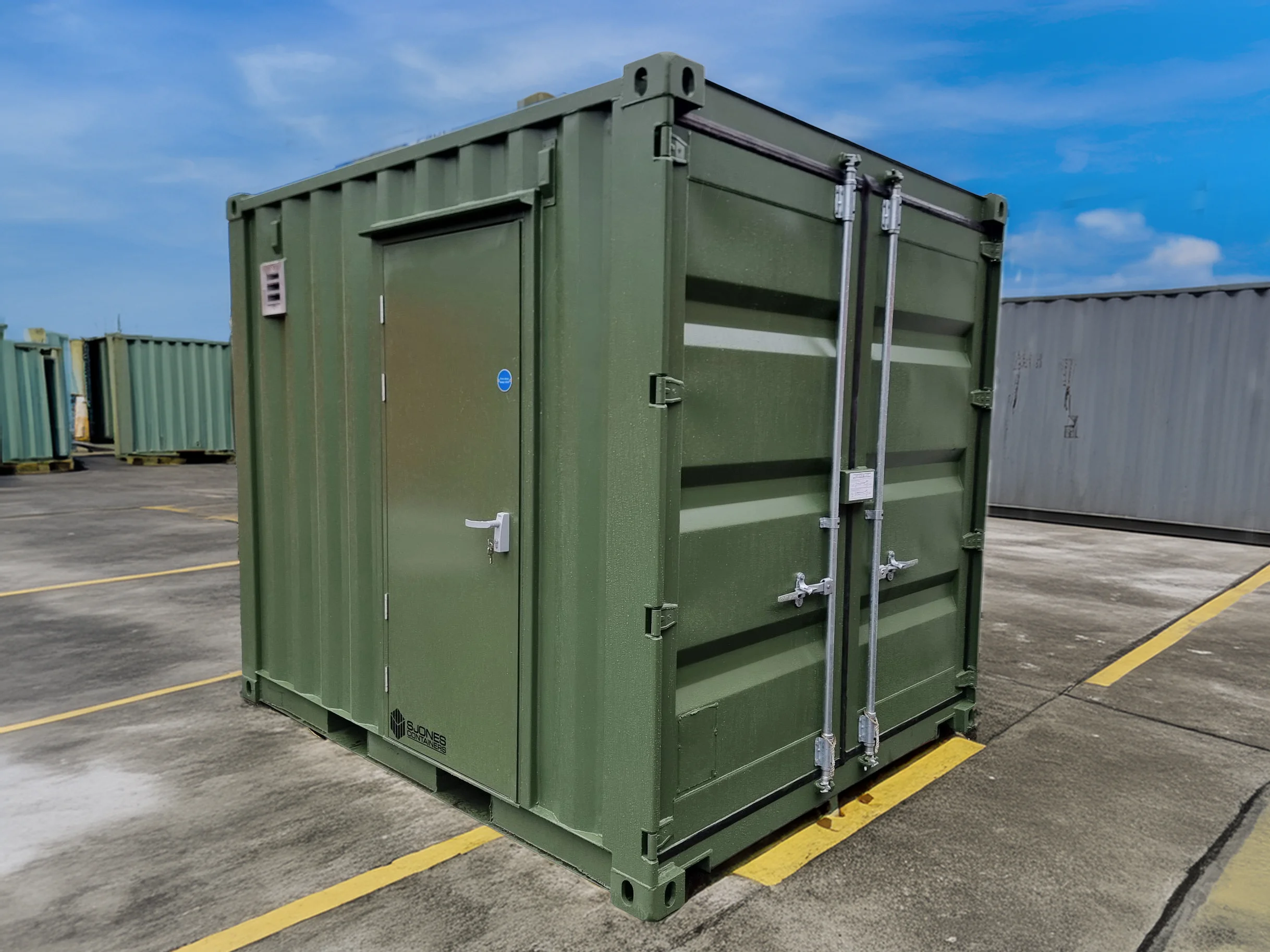 This image shows a compact, 10-foot metal container spray-painted in dark olive green. The container is situated outdoors on a paved area, marked with yellow parking lines, under a clear blue sky. The container features a secured personnel door on the left side with a handle and lock, and double cargo doors on the front. The surroundings include other similarly sized containers, suggesting a storage or industrial facility.