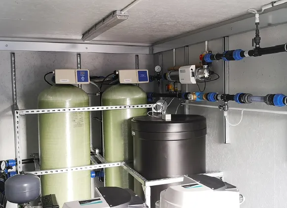 Interior of a containerised water treatment system featuring cylindrical filtration tanks, a black water tank, and an array of connected pipes, valves, and electronic control units mounted on the wall.