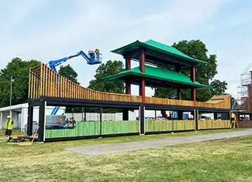 Midway through construction, the Sakura event installation features a two-tiered pagoda-style structure with green roofs and red vertical supports atop a base made from black shipping containers. Workers on a cherry picker install decorative timber slats along the top edge. The setup is taking place on a grassy field with trees in the background.