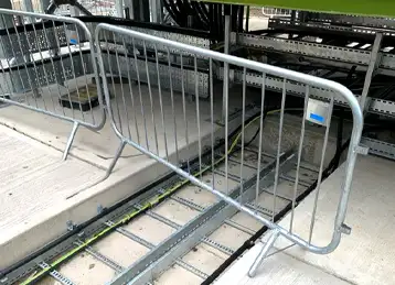 A temporary metal safety barrier positioned in front of exposed cable trays running along the ground at an industrial site. The cables are protected by metal trunking, and the area is partially raised with a concrete platform and metal framework above.