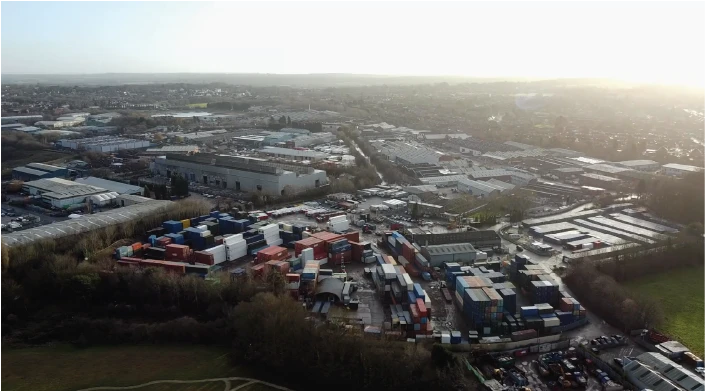 An aerial view of an industrial area with large warehouses, buildings, and a shipping container yard filled with colorful stacked containers. The surrounding landscape includes trees, roads, and an open field.
